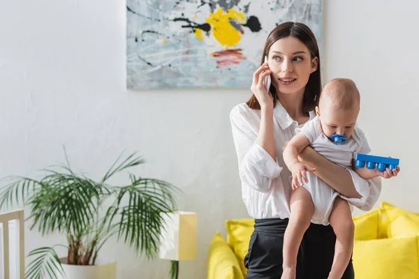 Woman talking on cellphone while working at home with toddler kid in hands — Stock Photo