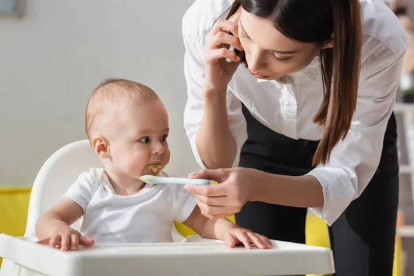 Woman talking on mobile phone while feeding toddler son with puree — Stock Photo