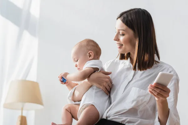 Sonriente mujer sosteniendo el teléfono móvil y niño pequeño en casa — Stock Photo