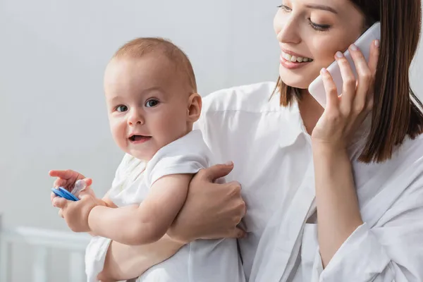 Усміхнена жінка розмовляє на мобільному телефоні, тримаючи малюка вдома — стокове фото