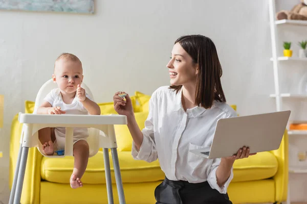 Niño en silla de bebé mostrando el pulgar hacia arriba cerca de mamá feliz con cuchara y portátil - foto de stock