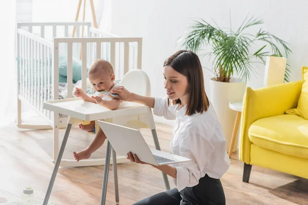 Femme brune assise sur le sol avec un ordinateur portable tout en nourrissant son fils dans une chaise bébé — Photo de stock