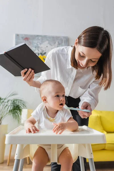 Cheerful woman with notebook feeding little son with puree at home — Stock Photo