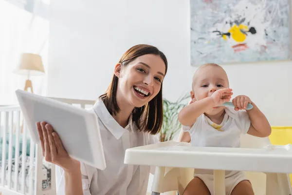 Fröhliche Frau mit digitalem Tablet blickt in Kamera, während sie Kleinkind-Sohn im Babystuhl füttert — Stockfoto