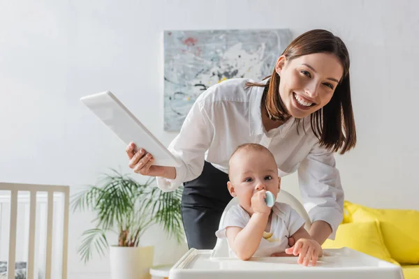 Donna allegra con tablet digitale guardando la macchina fotografica vicino a piccolo figlio seduto sulla sedia del bambino con cucchiaio — Foto stock