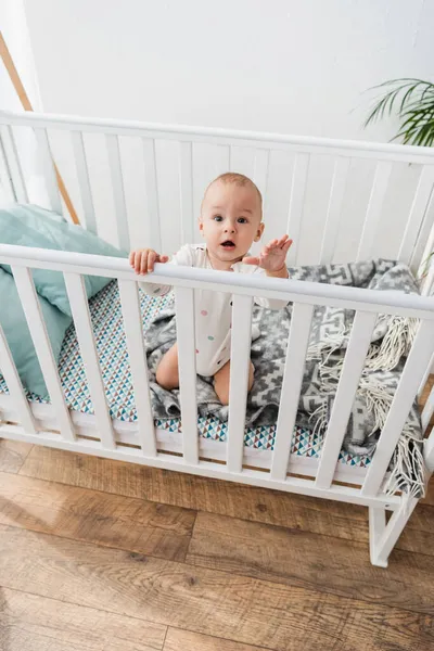 Aus der Vogelperspektive betrachtet, wie ein kleiner Junge in die Kamera schaut, während er im Kinderbett sitzt — Stockfoto