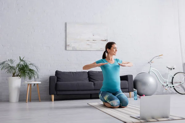 Mujer embarazada feliz en ropa deportiva ejercicio cerca de la computadora portátil en la sala de estar - foto de stock