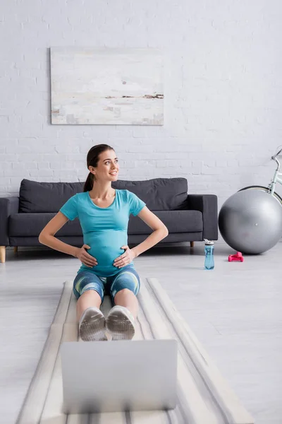 Glückliche Schwangere in Sportbekleidung, die in der Nähe von Laptop auf Fitnessmatte trainiert — Stockfoto