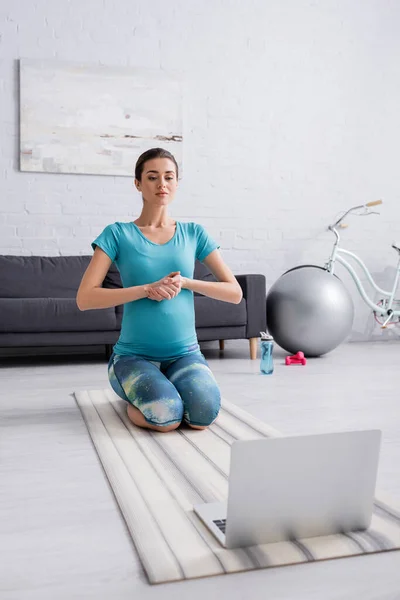 Mujer embarazada joven en ropa deportiva haciendo ejercicio cerca de la computadora portátil en la sala de estar - foto de stock