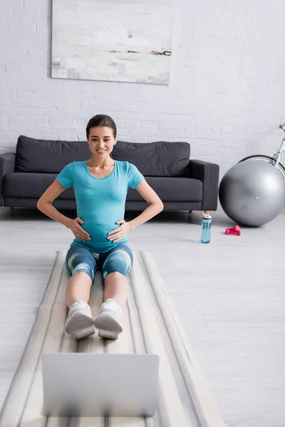 Mujer embarazada sonriente en ropa deportiva ejercicio cerca de la computadora portátil en la sala de estar — Stock Photo