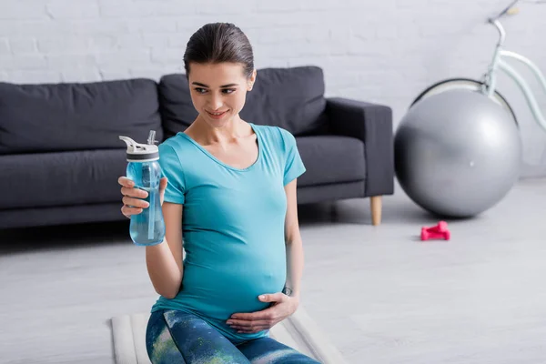 Счастливая беременная женщина смотрит на спортивную бутылку в гостиной — стоковое фото