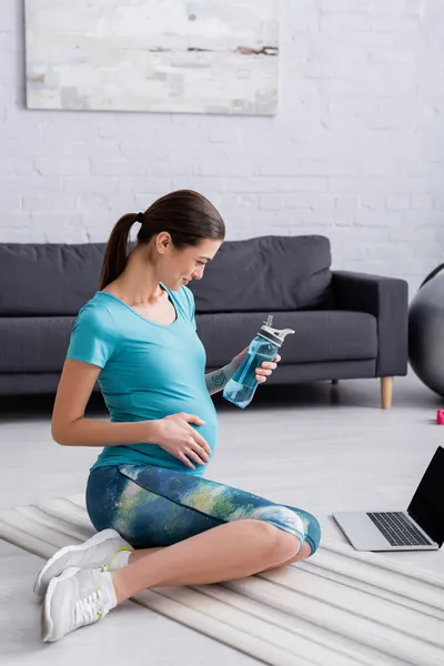 Улыбающаяся беременная женщина смотрит на ноутбук, держа в руках спортивную бутылку — стоковое фото