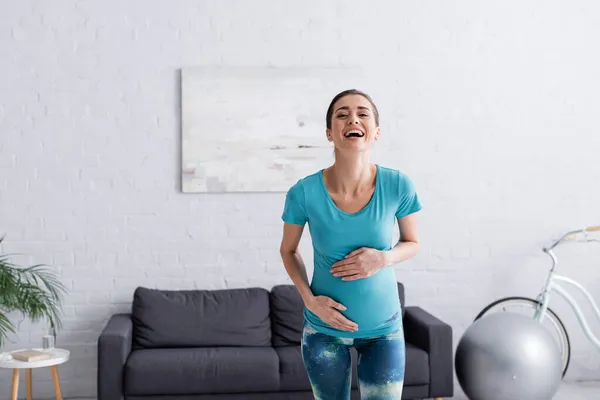 Alegre desportista grávida tocando barriga e rindo na sala de estar — Fotografia de Stock