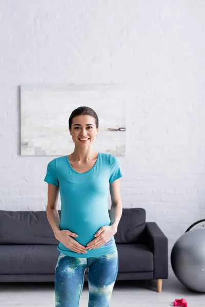 Deportista embarazada positiva tocando el vientre en la sala de estar - foto de stock