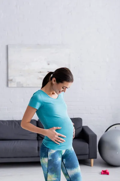 Cheerful pregnant sportswoman touching belly in living room — Stock Photo