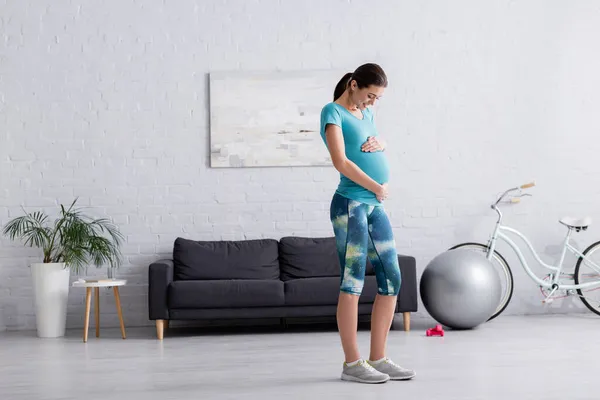 Longitud completa de la mujer embarazada feliz tocando vientre en la sala de estar - foto de stock