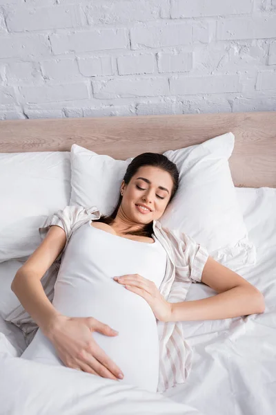 Mujer embarazada despierta tocando el vientre mientras está acostada en la cama - foto de stock