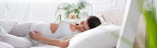 Mujer embarazada complacida tocando el vientre mientras está acostada en la cama, pancarta - foto de stock