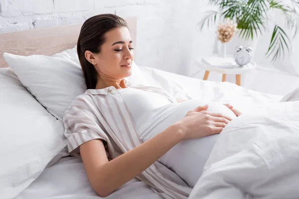 Mujer embarazada complacida tocando el vientre mientras está acostada en la cama - foto de stock