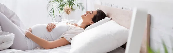 Вид збоку щаслива молода вагітна жінка торкається живота, лежачи в спальні, банер — стокове фото