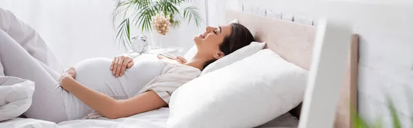 Side view of happy young pregnant woman touching belly while lying in bedroom, banner — Stock Photo