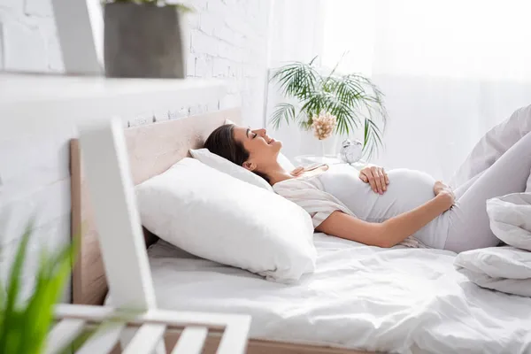 Vista laterale di allegra giovane donna incinta che tocca la pancia mentre si trova in camera da letto — Foto stock