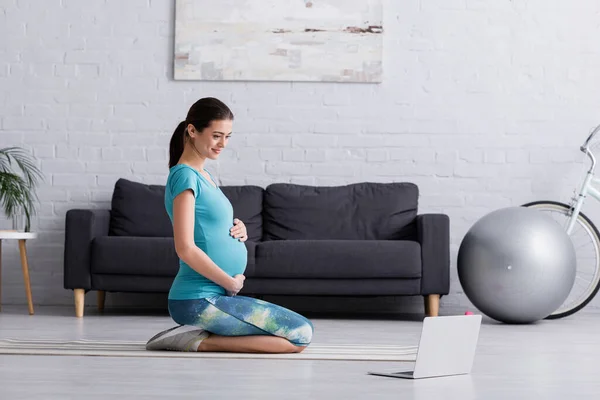 Mujer embarazada feliz en ropa deportiva trabajando cerca de la computadora portátil en la estera de fitness - foto de stock