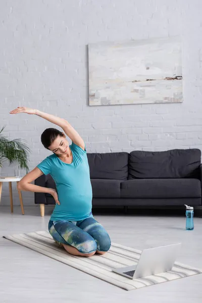 Cheerful pregnant woman in sportswear stretching with hand on hip while looking at laptop — Stock Photo