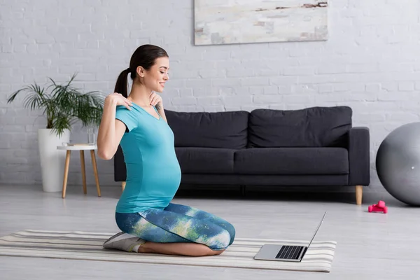Улыбающаяся беременная женщина в спортивной одежде тренируется, глядя на ноутбук — стоковое фото