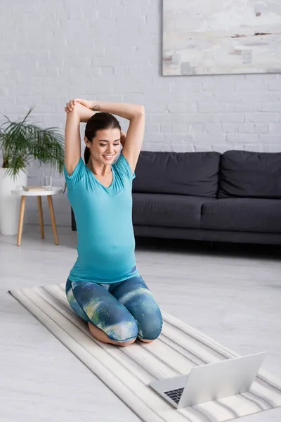 Tattooed pregnant sportswoman in sportswear working out while smiling near laptop — Stock Photo
