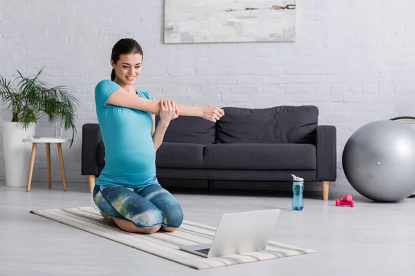 Happy pregnant woman in sportswear stretching while looking at laptop — Stock Photo