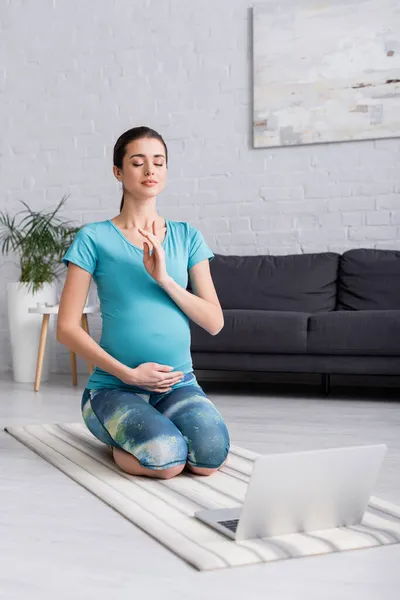 Joven embarazada con los ojos cerrados meditando en la sala de estar - foto de stock