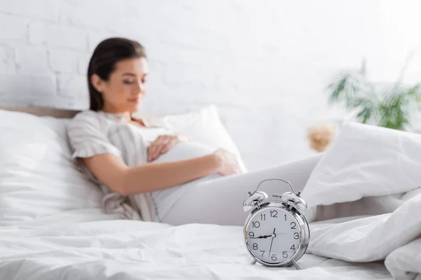 Ретро будильник на кровати рядом с размытой беременной женщиной — стоковое фото
