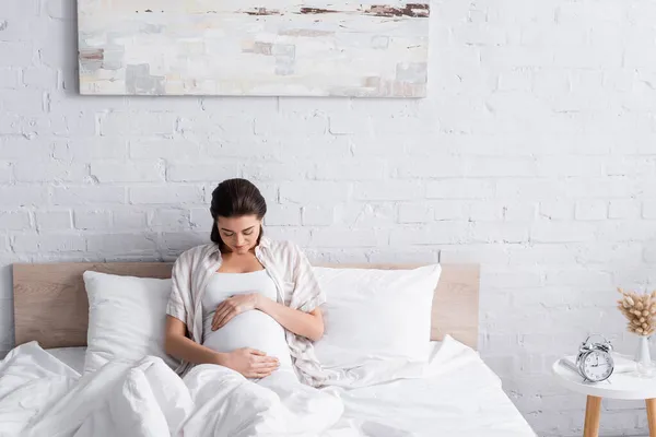 Mujer joven feliz y embarazada tocando el vientre en el dormitorio - foto de stock