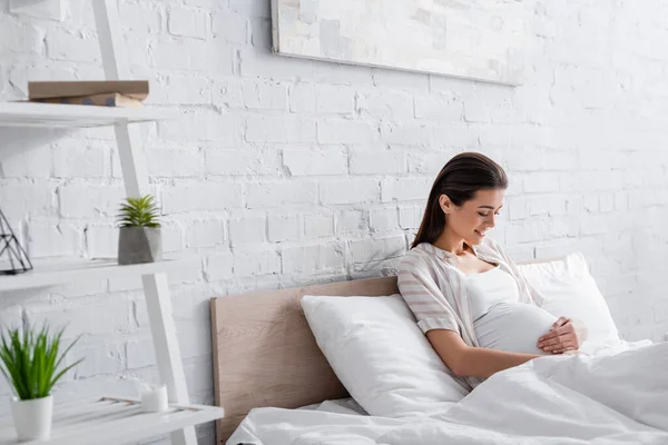 Happy pregnant woman looking at belly in bedroom — Stock Photo