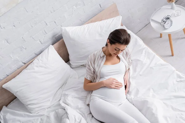 Vista de ángulo alto de la mujer embarazada sonriente mirando el vientre en el dormitorio - foto de stock