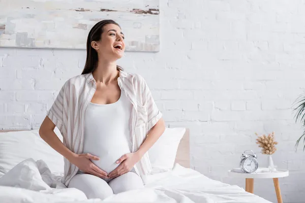 Glücklich schwangere Frau lacht und berührt Bauch im Schlafzimmer — Stockfoto