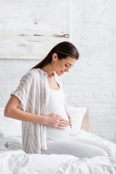 Happy pregnant woman looking and touching belly in bedroom — Stock Photo