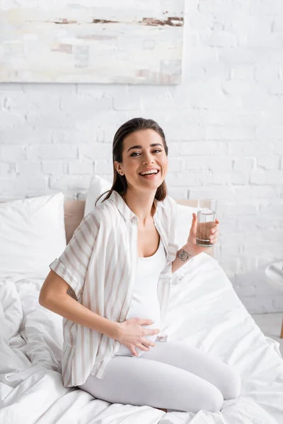 Mujer embarazada feliz sosteniendo vaso de agua en el dormitorio - foto de stock