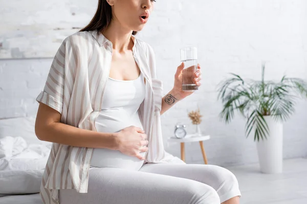 Vista recortada de la mujer embarazada sosteniendo un vaso de agua mientras siente dolor abdominal - foto de stock
