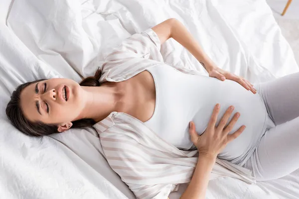 Top view of young pregnant woman feeling cramp while lying on bed — Stock Photo