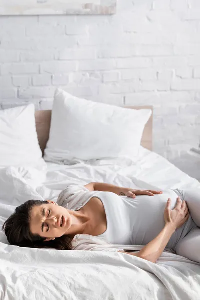 Молодая беременная женщина с закрытыми глазами страдает от спазмов во время лежания на кровати — стоковое фото