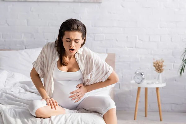 Young pregnant woman with closed eyes suffering from cramp while sitting on bed — Stock Photo