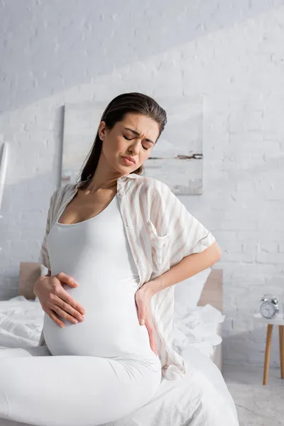 Brunette pregnant woman with closed eyes feeling abdominal pain in bedroom — Stock Photo