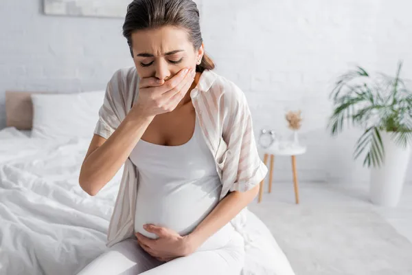 Mujer embarazada joven sintiendo náuseas y cubriendo la boca en el dormitorio - foto de stock