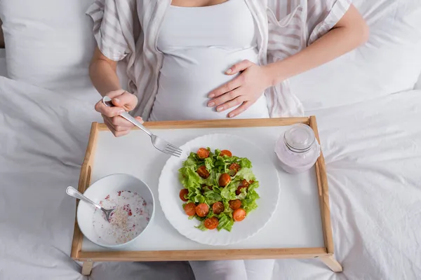 Обрезанный вид беременной женщины с вилкой рядом с едой на подносе — стоковое фото