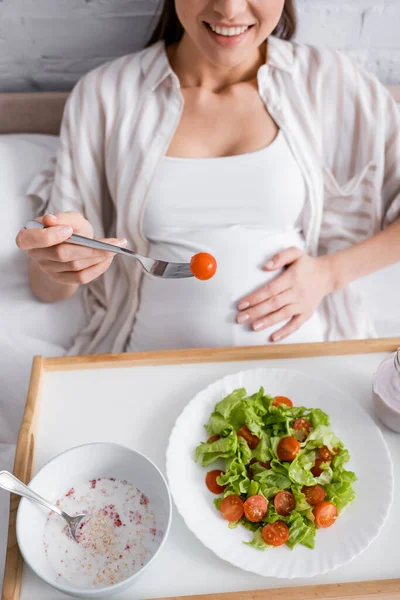 Vista recortada de la mujer embarazada feliz sosteniendo tenedor con tomate cereza cerca de la comida en bandeja - foto de stock