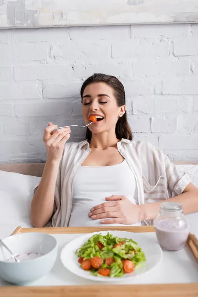 Mujer embarazada feliz sosteniendo tenedor con tomate cereza cerca de la comida en bandeja - foto de stock