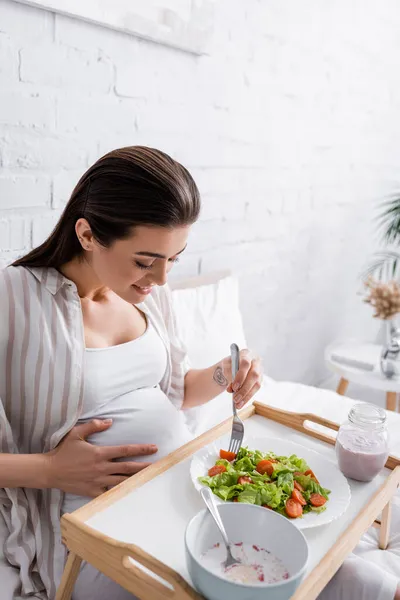 Татуированная и счастливая беременная женщина с вилкой рядом с салатом на подносе — стоковое фото