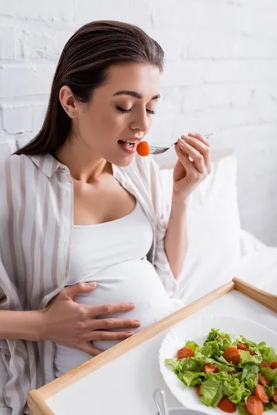 Брюнетка беременная женщина держит вилку во время еды помидор черри — стоковое фото